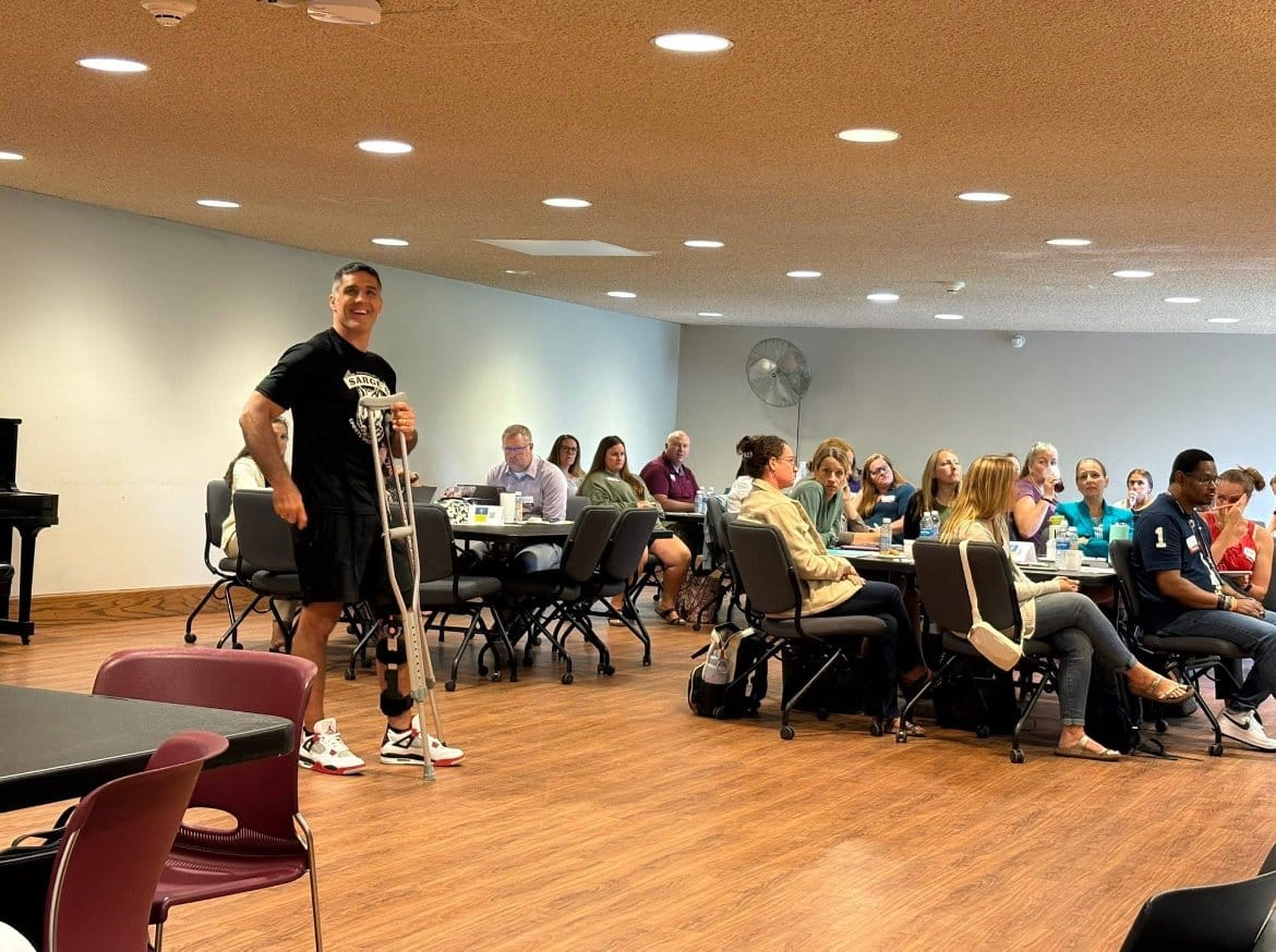 Chris Sargent on crutches presenting to a room of adults seated at tables